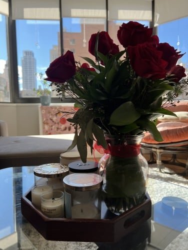 A vase of red roses on a glass table. backlit by large windows showing a blue sky and buildings.