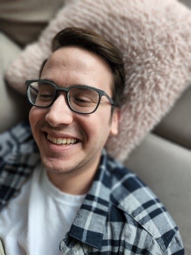 Man wearing glasses smiling and laying against a pink fluffy pillow, taken with a blurred background effect.