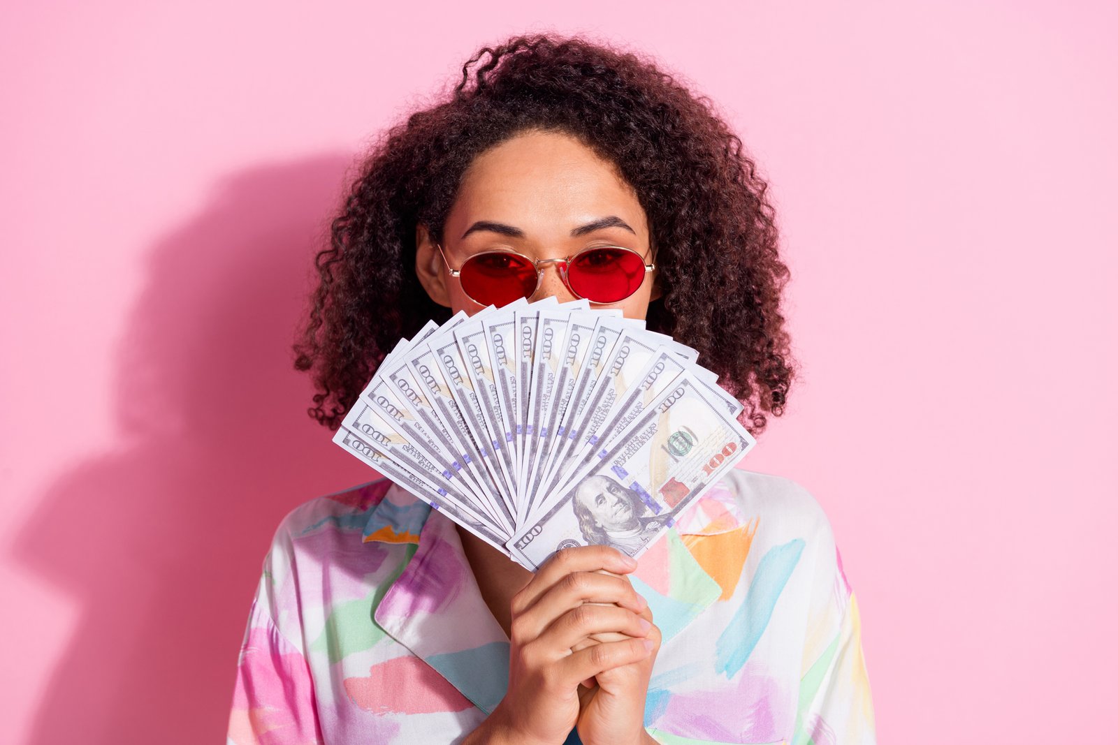 A woman folds a fan of $100 bills in front of her face in front of a pink background.