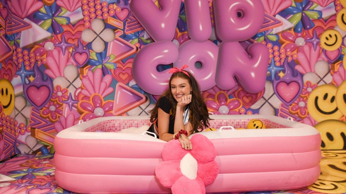 woman in pink inflatable tub with daisy posing for camera with balloons reading VidCon behind her