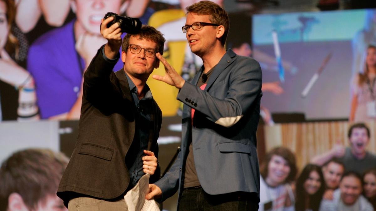 two men with glasses speaking in front of a crowd and taking a selfie with a SLR camera
