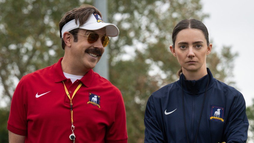 A coach with a mustache, visor and sunglasses smiles at a serious looking woman next to him.